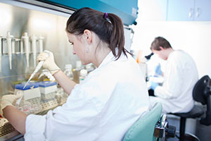In foreground, a young woman in white lab coat and wearing latex gloves uses syringe to extract specimen from a bottle while in background a young man in similar lab coat peers into microscope