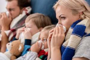 Close-up shot of sick young family--mom, dad, and preschool-age boy and girl--blowing noses with tissues together while lying in bed