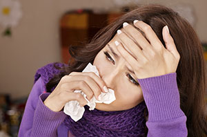 Young woman looks miserable, holds tissue to her nose with one hand and places the other on her forehead