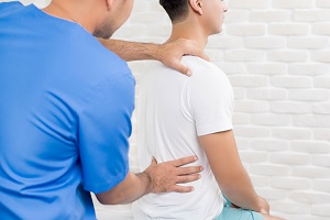 Rear view of male medical practitioner examining a young adult male for back pain, with one hand on the patient's shoulder and the other mid-torso