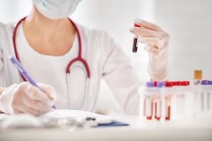 Close-up of a medical practitioner holding a blood test tube and making notes. The practitioner is sitting at a table in a lab with a row of sample vials in front of her.