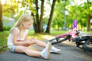 Cute little girl sitting on the ground, clutching her knee, which has a large cut after she fell off her bike at a park.