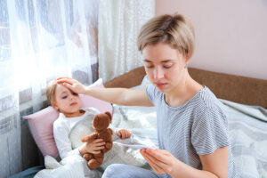 Mother measuring temperature of her sick child who is laying in bed with teddy bear